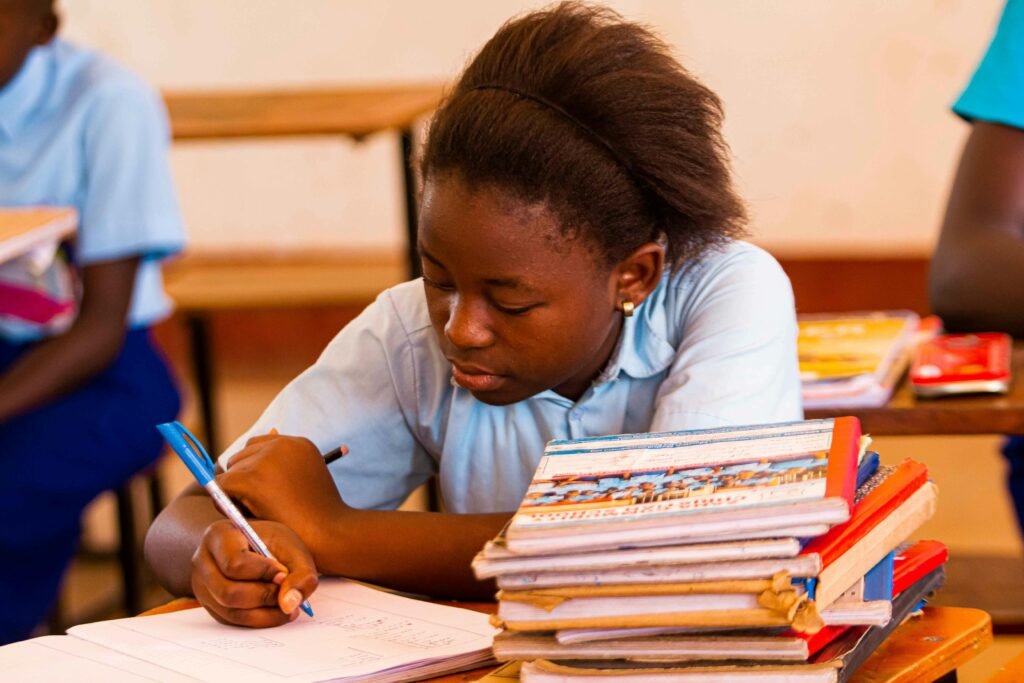 A schoolgirl in uniform writing in a notebook surrounded by books in a classroom setting.
